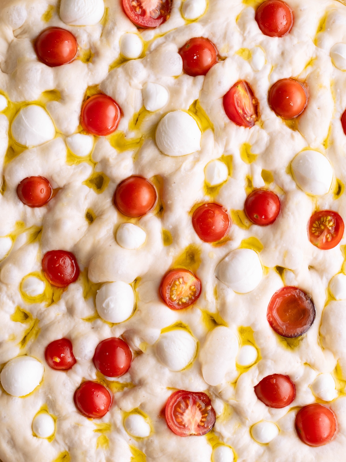 Bubbly focaccia bread dough dimpled with cherry tomatoes, basil flower oil, and mozzarella balls
