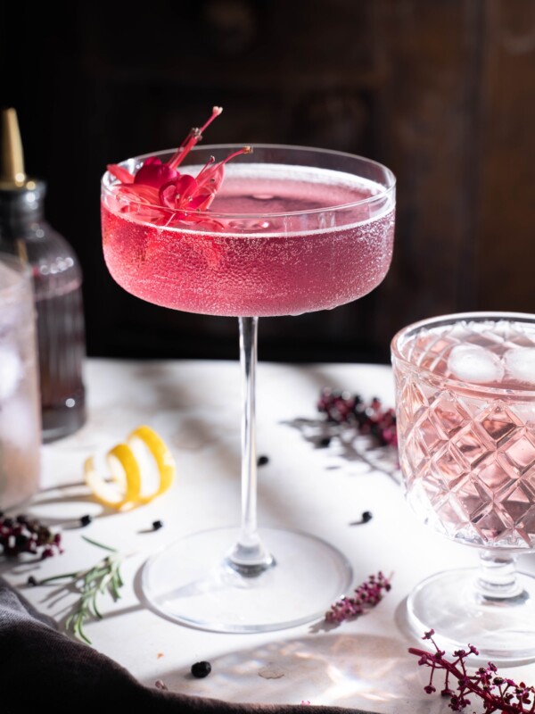 A beautifully pink elderberry cocktail served in a tall coupe glass with an edible flower garnish