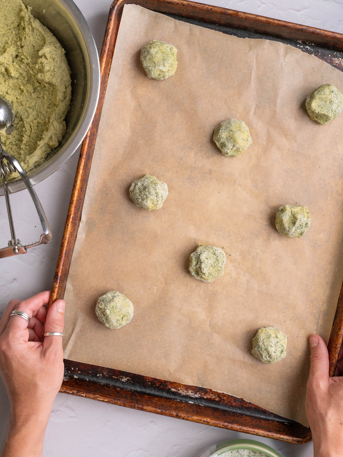 Cookie dough balls ready to bake on a sheet pan, next to a bowl of cookie dough